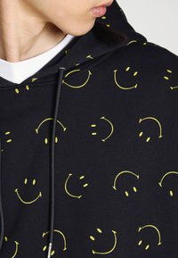 Black hoodie featuring a repeating pattern of yellow smiley faces and arrows. Material appears soft with a textured finish and adjustable drawstrings.