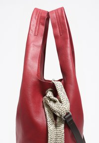 Rdeča usnjena torba z gladko teksturo, vozlana bež in črna tekstilna naramnica ter dva navpična ročaja za nošenje.