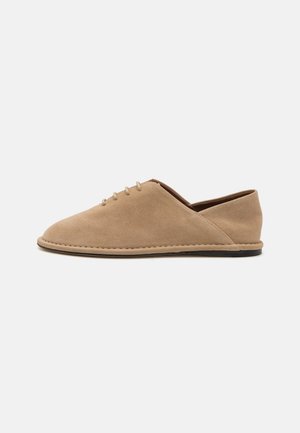 Beige suède schoenen met een laag profiel, ronde neus en vijf veterogen. Stikseldetails langs de rand en een donkere zool.