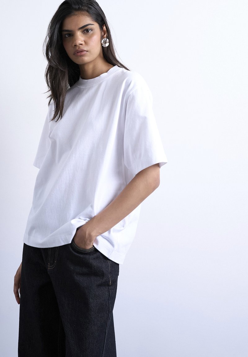 Νέα γυναίκα με μακριά σκούρα μαλλιά φοράει υπερμεγέθη λευκό μπλουζάκι και μαύρο τζιν, ποζάροντας με το ένα χέρι στην τσέπη μπροστά σε απλό ανοιχτό φόντο.