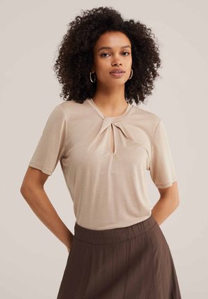 Femme aux cheveux bouclés portant des boucles d’oreilles créoles dorées, un top beige à manches courtes avec un détail torsadé à l’avant, et une jupe marron taille haute, posant les mains dans le dos.