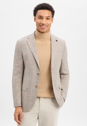 Junger Mann lächelt, trägt einen beigefarbenen Rollkragenpullover, einen karierten Blazer und hellfarbene Hosen, steht vor einem schlichten hellen Hintergrund.