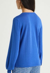 Pull en maille bleu avec manches longues légèrement bouffantes et encolure ronde. Texture lisse, sans motifs apparents, porté avec un pantalon à motifs.