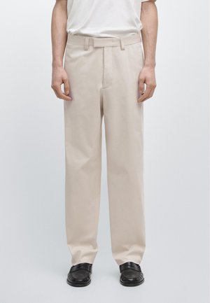 Mann trägt beige gerade geschnittene Hosen, weißes T-Shirt und schwarze Mokassins, steht vor einem schlichten hellen Hintergrund.