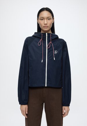 Vrouw met lang zwart haar die een marineblauwe capuchontrui draagt met een witte ritssluiting en rood-witte trekkoorden, gecombineerd met een bruine broek, staand tegen een effen achtergrond.