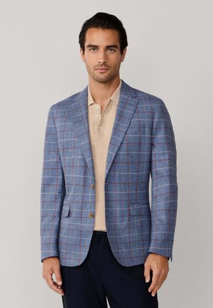 Man draagt een lichtblauw geruit colbert over een beige poloshirt en donkere broek, staand tegen een effen lichte achtergrond.