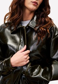 Veste aviateur en faux cuir vert avec un col montant, une fermeture à pression à l'avant et des poignets côtelés. La texture est brillante avec une finition lisse.