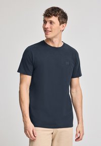 Granatowa bawełniana koszulka z krótkim rękawem, okrągłym dekoltem i małą kwadratową łatką z logo na piersi. Gładka tekstura, casualowy design.