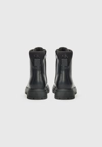 Botas de cuero negras hasta el tobillo con superficie lisa, cremallera lateral y suelas planas de goma. Presentan un logo en el talón y un collarín acolchado.