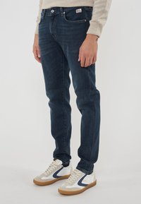 Uomo che indossa jeans blu scuro, un maglione beige e scarpe da ginnastica bianche con dettagli navy e beige, in piedi contro uno sfondo chiaro e semplice.
