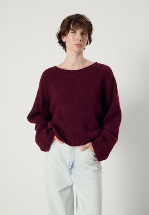 Person mit kurzen lockigen Haaren trägt einen locker sitzenden weinroten Pullover und hellblaue High-Waist-Jeans, steht vor einem schlichten weißen Hintergrund.