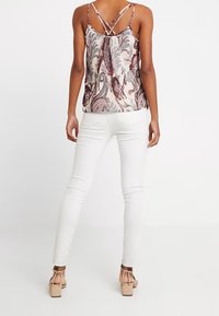 Kvinna iklädd ett mönstrat ärmlöst linne med korsade axelremmar, vita tajta jeans och beige höga klackar, stående med ryggen mot betraktaren.