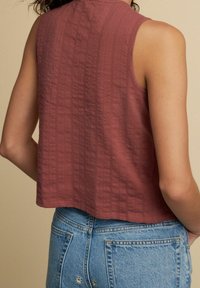 Rostfarbener ärmelloser Oberteil aus strukturiertem Stoff. Kurzes Design mit geradem Saum, kombiniert mit hellblauen High-Waist-Jeans.