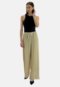 Schwarzes ärmelloses Oberteil kombiniert mit beigen plissierten weiten Hosen mit elastischem Bund und Kordelzug. Schwarze spitze Schuhe.