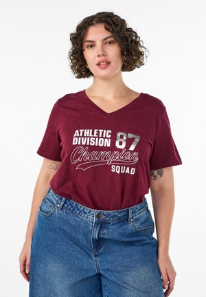 Kehaga naine, kellel on lokkis juuksed, riietatud burgundiapunasesse "Athletic Division 87 Champion Squad" T-särki ja kõrge vöökohaga sinistesse teksadesse, seisab valge tausta ees.
