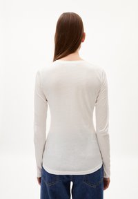Person mit langen braunen Haaren, der ein eng anliegendes weißes Langarmshirt und blaue Jeans trägt, steht mit dem Rücken zu einer einfarbigen Hintergrund.