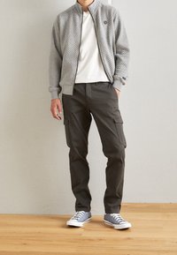 Giacca grigia trapuntata con zip sopra una camicia bianca, abbinata a pantaloni cargo verde scuro e sneakers grigie con suola bianca. In piedi su pavimento in legno.