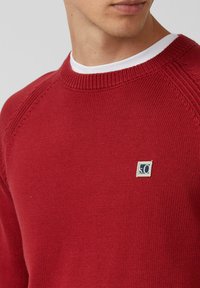 Mann trägt einen roten, gestrickten Rundhalsausschnitt-Pullover mit einem kleinen, marineblauen und weißen quadratischen Logo auf der Brust über einem weißen T-Shirt.