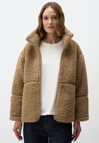 Beige nepbontjas met een rits aan de voorkant, hoge kraag en brede mouwen. Getextureerd oppervlak met een pluche gevoel, gecombineerd met een witte top.