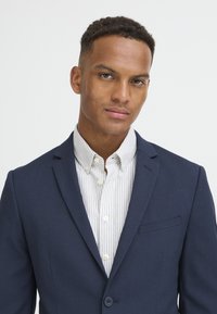 Sötétkék zakó, amely testre szabott szabású, kipróbált hajtókával és mellzsebkendővel rendelkezik. Alatta egy világos csíkos gombos ing látható.