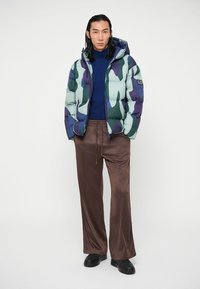 Homem com uma jaqueta puffer azul camuflada, um suéter de gola alta azul-marinho, calças largas castanhas e sapatos pretos, de pé contra um fundo branco.