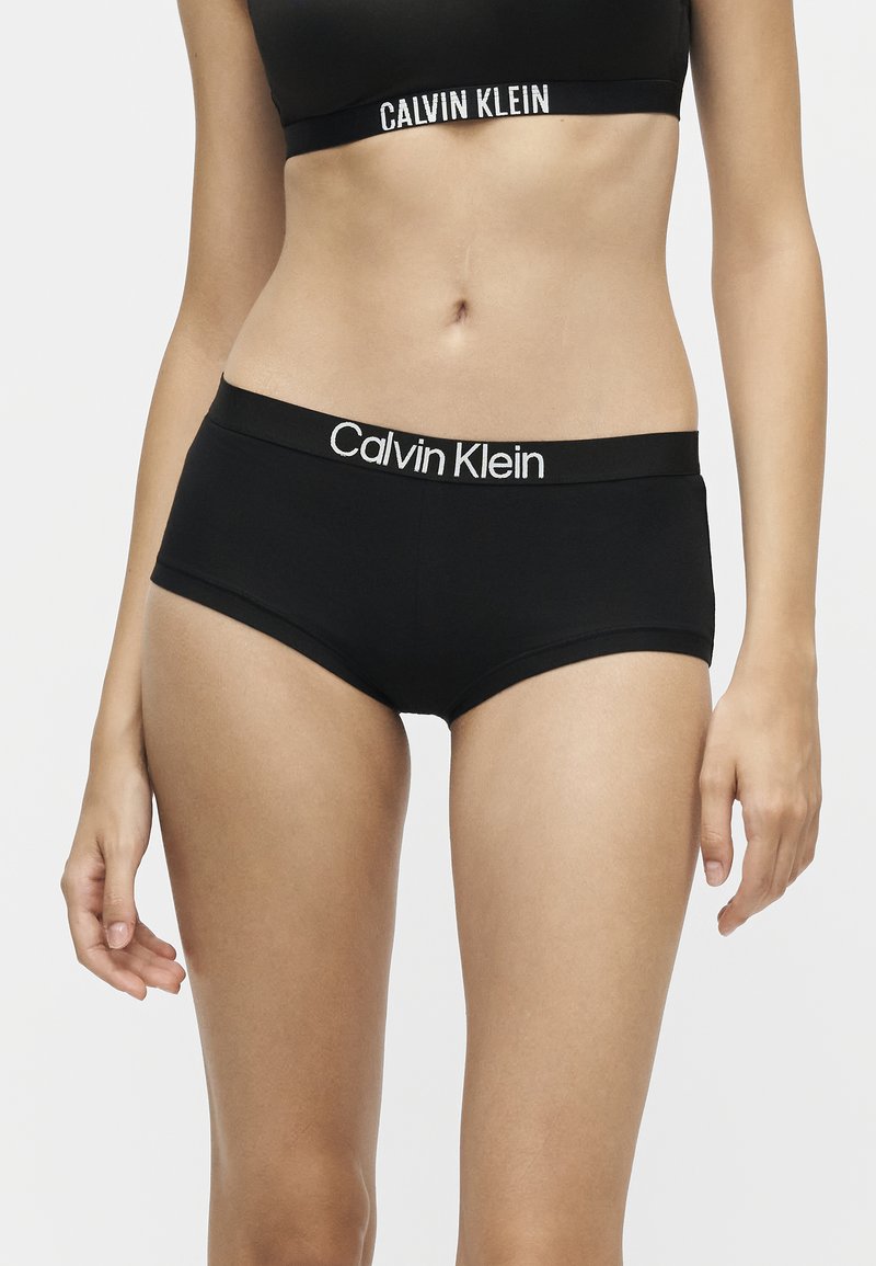 Γυναίκα με μαύρο σετ εσωρούχων Calvin Klein, που δείχνει τη μέση και το άνω μέρος των μηρών σε απλό φωτεινό φόντο.