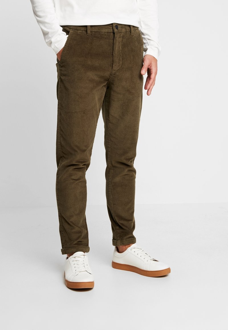 Pantalon en velours côtelé marron à coupe slim, avec poches, passants de ceinture et fermeture à bouton, associé à des baskets blanches.