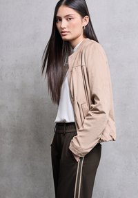 Giacca bomber beige con polsini a costine, chiusura con zip e tasche laterali, indossata sopra una camicia bianca e pantaloni marroni con strisce laterali.