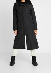 Manteau long noir imperméable avec capuche, fermeture éclair sur le devant, deux grandes poches et poignets réglables. Porté avec un pantalon neutre et des bottes noires.