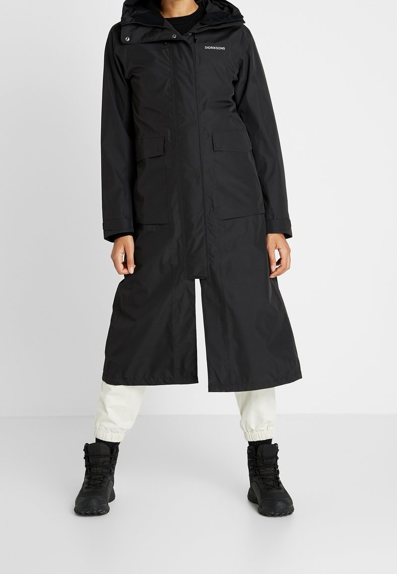 Manteau long noir imperméable avec capuche, fermeture éclair sur le devant, deux grandes poches et poignets réglables. Porté avec un pantalon neutre et des bottes noires.