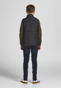 Zwarte puffervest met horizontale stiksels, groene mouwen, donkere jeans en witte schoenen met donkere accenten, gezien vanaf de achterkant.