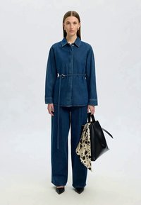 Mulher vestindo uma jaqueta de ganga azul-escura e calças, segurando uma bolsa preta com um lenço estampado, de pé contra um fundo claro.