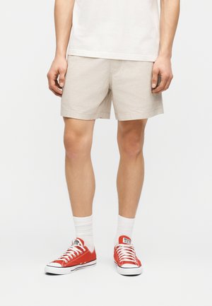 Mann trägt beige legere Shorts, weiße Socken und leuchtend rote niedrige Sneakers, steht vor einfarbigem Hintergrund.