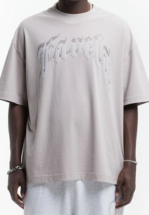 Czarna osoba ubrana w luźny jasnoszary T-shirt z perforowanym gotyckim tekstem oraz szare szorty, uzupełnione srebrnymi bransoletkami i perłowym naszyjnikiem.