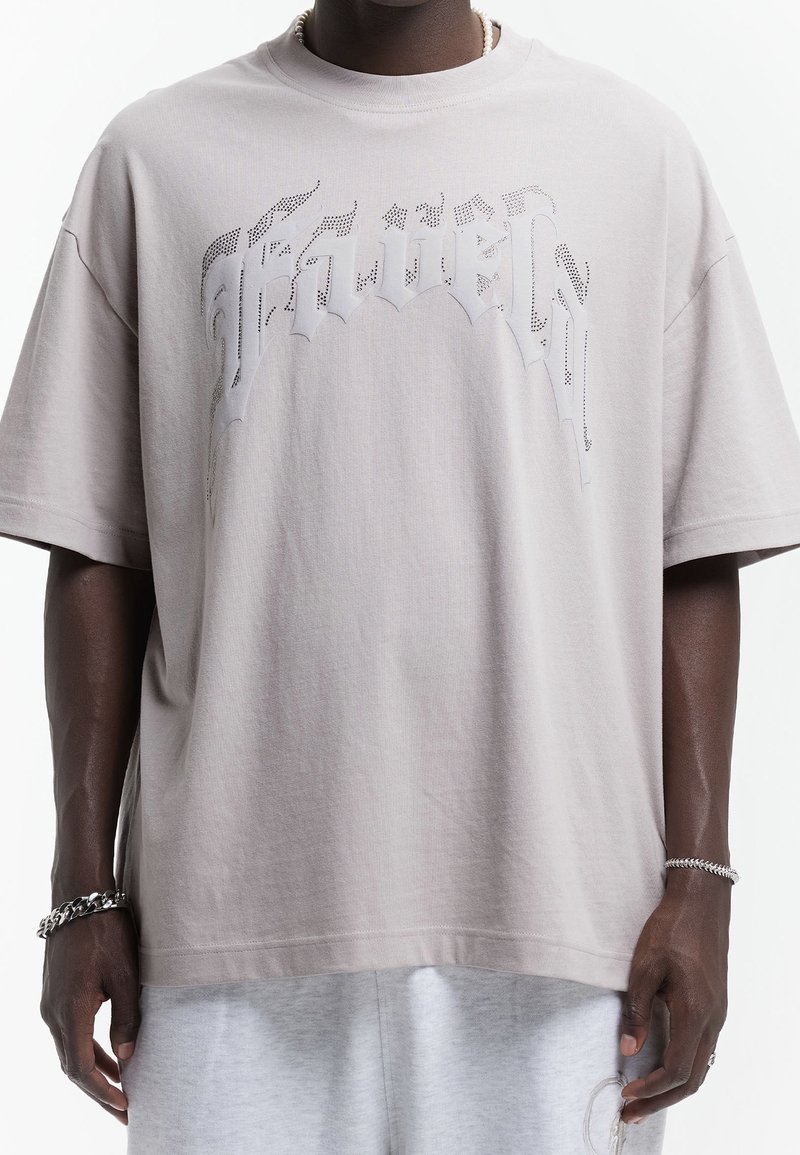 Personne noire portant un t-shirt ample gris clair avec un texte gothique perforé et un short gris, accessoirisée de bracelets argentés et d'un collier de perles.