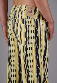 Man wearing yellow, black, and white patterned jeans with two back pockets and a leather brand label on the waistband.