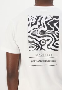 Balta īsām piedurknēm t-krekls ar melnbaltu abstraktu rakstu un "Columbia" logo ar tekstu: "Kopš 1938. gada, Portlenda, Oregona, ASV."