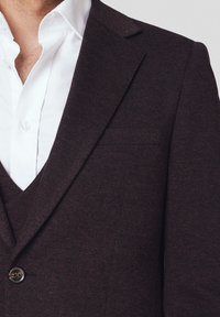 Ciemnobrązowy blazer o fakturze z pojedynczym zapięciem na guzik i klapami w kształcie notcha, noszony na białej koszuli z kołnierzykiem.