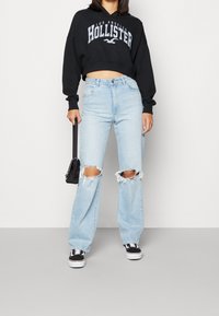 Felpa nera corta con grafica "Hollister", jeans a gamba larga azzurri chiari con ginocchia strappate e sneakers nere con suola bianca. Porta una piccola borsa nera.