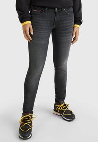 Jeans negros ajustados con textura desgastada y costuras sutiles. Llevados con zapatillas negras que tienen cordones amarillos y una suela resistente.