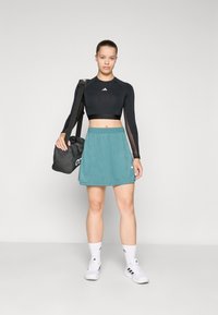 Fekete, hosszú ujjú crop top, türkiz színű sport szoknya, fehér zoknik és fehér tornacipő. A modell egy fekete táskát tart, amelyen jól látható a logó.