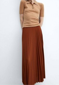Persona vestida con una camiseta polo de manga corta de canalé en tono marrón claro y una falda midi plisada de cintura alta en color naranja quemado, contra un fondo liso.