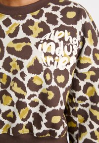 Mikina so vzorom leopardích skvrn v hnedých, krémových a olivových farbách s bielym nápisom "alpha industries". Má rebrovaný výstrih a manžety. Mäkká štruktúra tkaniny.