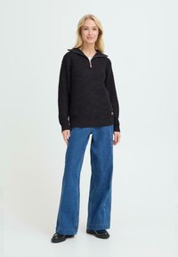 Svart zip-neck genser med strukturert strikk, sammen med vide blå jeans. Modellen står mot en lys bakgrunn og viser fram antrekket.
