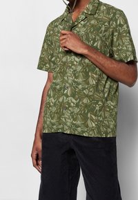 Uomo con camicia verde a maniche corte con stampa botanica a foglie, che tiene una tracolla marrone vicino al colletto, abbinata a pantaloni neri.