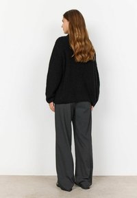 Un maglione oversize lavorato a maglia nero con un alto collo, abbinato a pantaloni grigi larghi. I capelli sono lunghi e mossi nella vista posteriore.