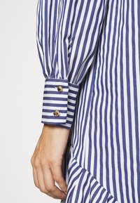 Bras gauche portant une chemise à manches longues rayée verticalement bleu et blanc avec un poignet fixé par deux boutons dorés.