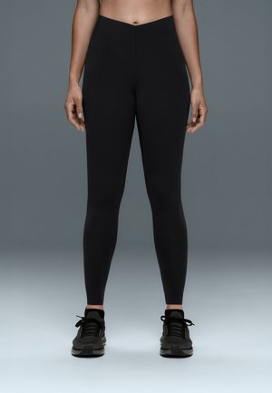 Femme portant un legging noir taille haute et des chaussures de sport noires, debout devant un fond gris uni, les mains détendues le long du corps.
