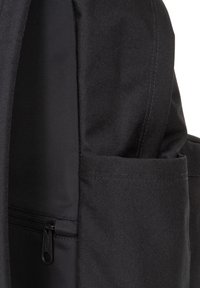 Mochila de tela negra con una superficie texturizada, que incluye un bolsillo con cremallera en el lateral y un bolsillo abierto. Costuras duraderas visibles a lo largo de las uniones.