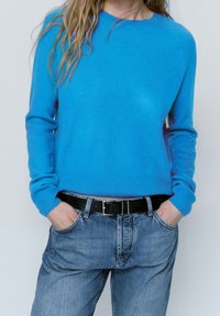 Maglione blu a maglia con scollatura rotonda, polsini a costine, abbinato a jeans in denim chiaro e una cintura nera. Tessuto morbido e vestibilità rilassata.